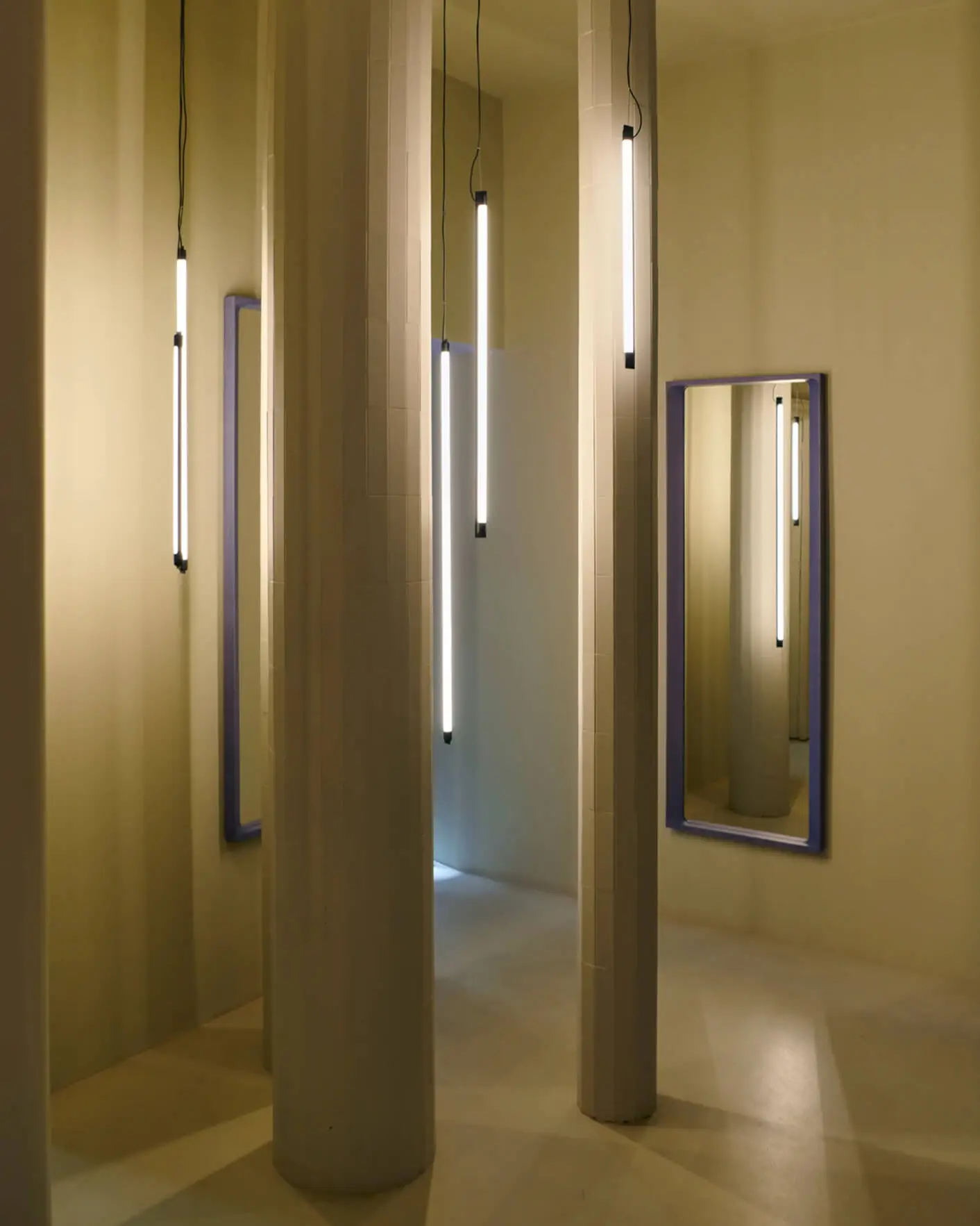 Minimalist interior with vertical linear pendant hanging lights, hexagonal columns all in beige with two mirrors on opposite walls with thick sculptural timber frames in bright purple finish.